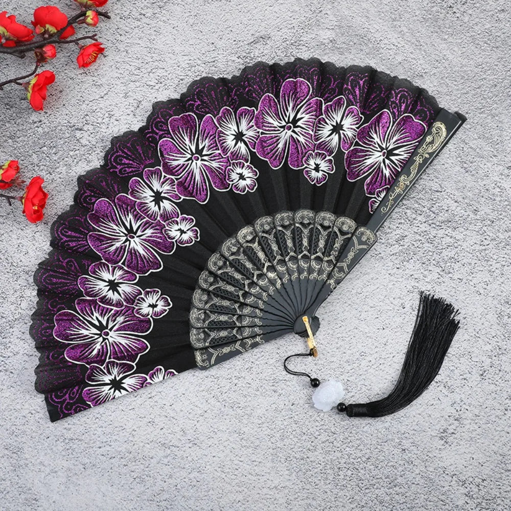 A dark purple and white floral antique folding fan with a black background and a black tassel.