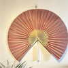 Handwoven bamboo wall fan decor with a reddish-brown color and a silver tassel mounted on a light wall.