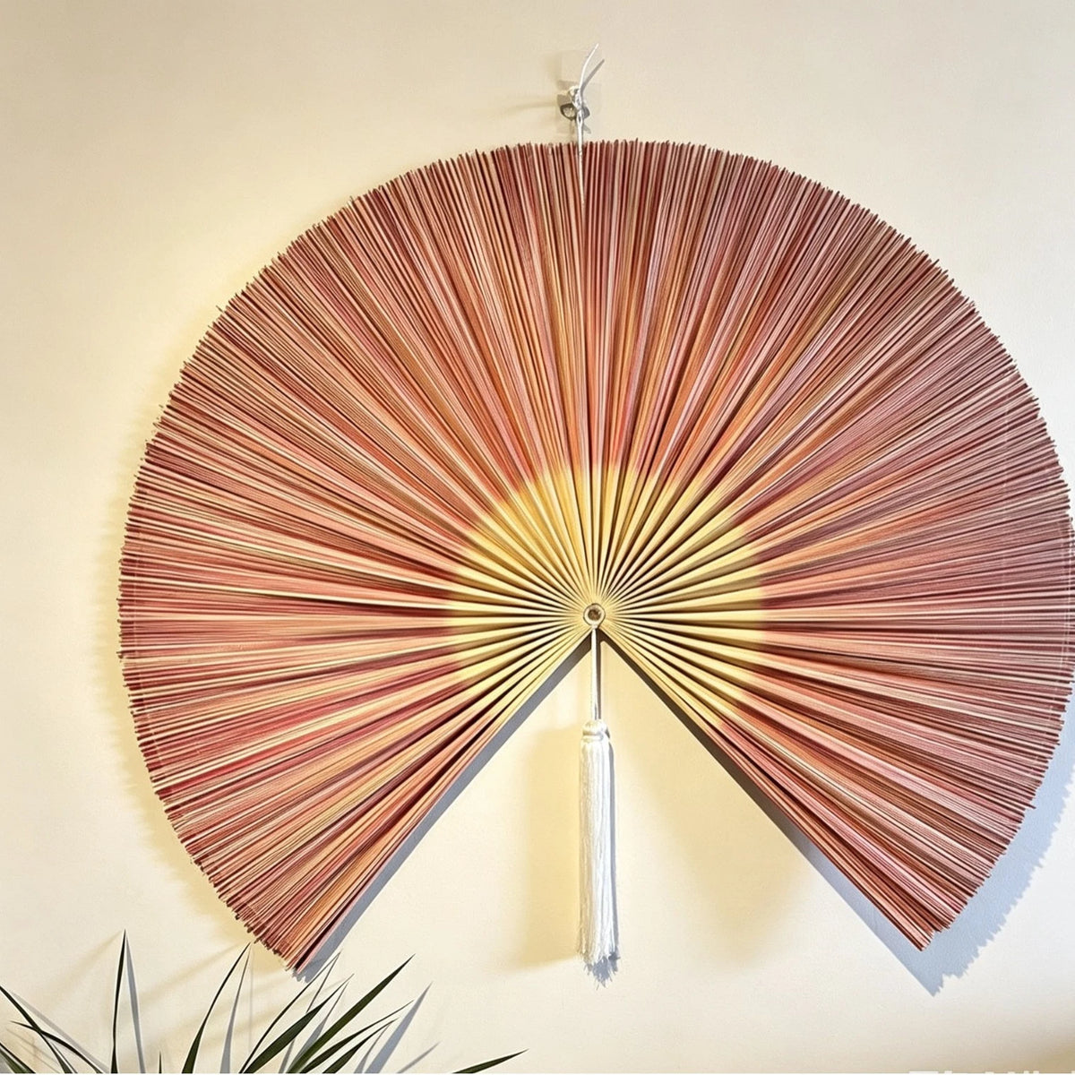 Handwoven bamboo wall fan decor with a reddish-brown color and a silver tassel mounted on a light wall.
