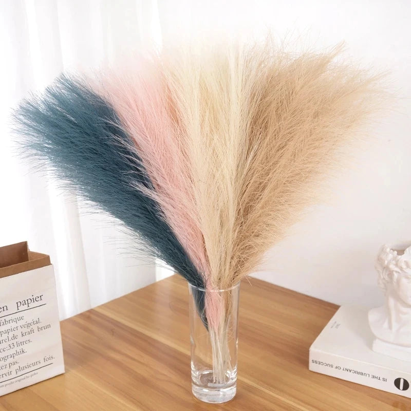 Un ramo de pampas artificiales en tonos azules, rosas y beige en un jarrón de vidrio transparente.