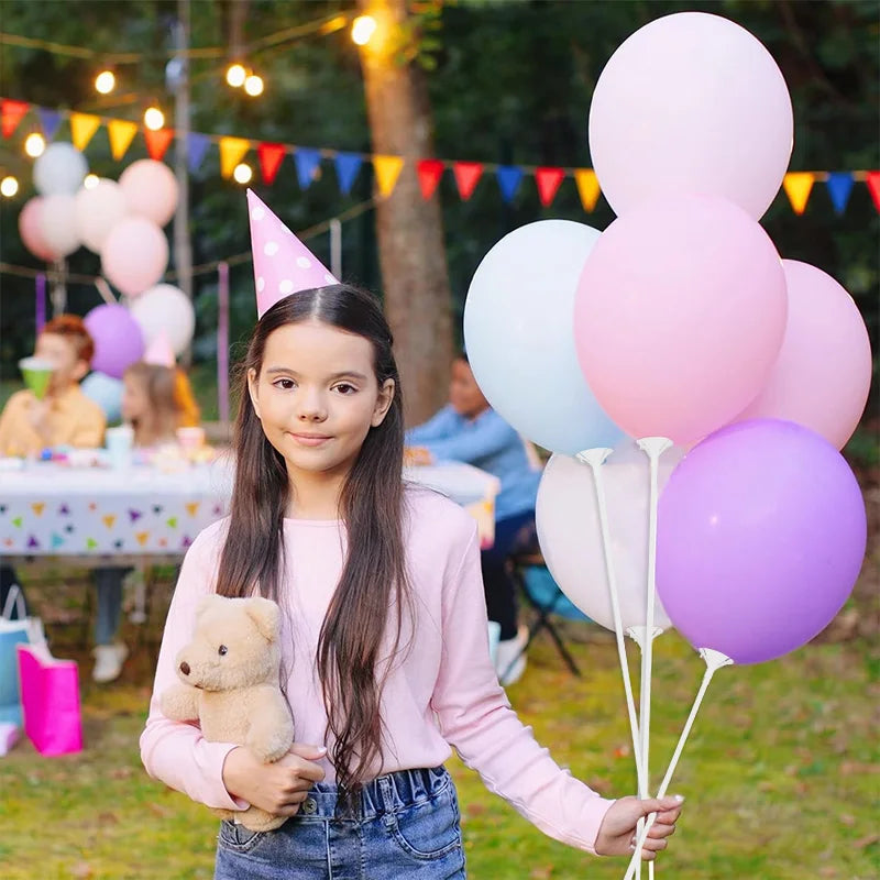 A girl holding a teddy bear stands in a grassy outdoor setting with pink and purple balloons.