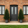 A dark gray front door flanked by two tall, conical artificial cedar topiary trees against a brick wall.