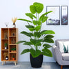 A large, lush artificial green tree with broad leaves sits in a black pot on a floor next to wooden shelving and framed art.