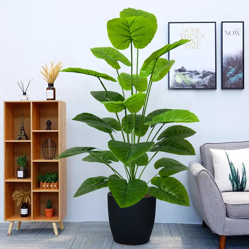A large, lush artificial green tree with broad leaves sits in a black pot on a floor next to wooden shelving and framed art.