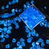 A pile of bright blue crystal ice cube acrylic stones displayed in a square glass container on black velvet fabric.