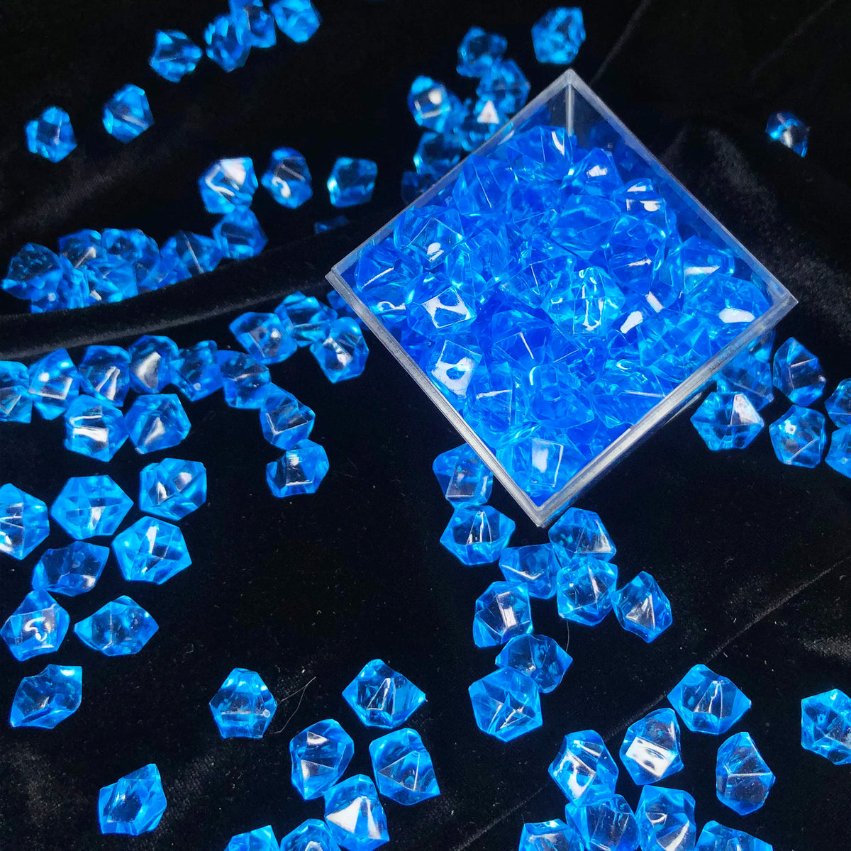 A pile of bright blue crystal ice cube acrylic stones displayed in a square glass container on black velvet fabric.