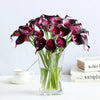 A bouquet of six artificial purple calla lilies in a glass vase on a white surface.