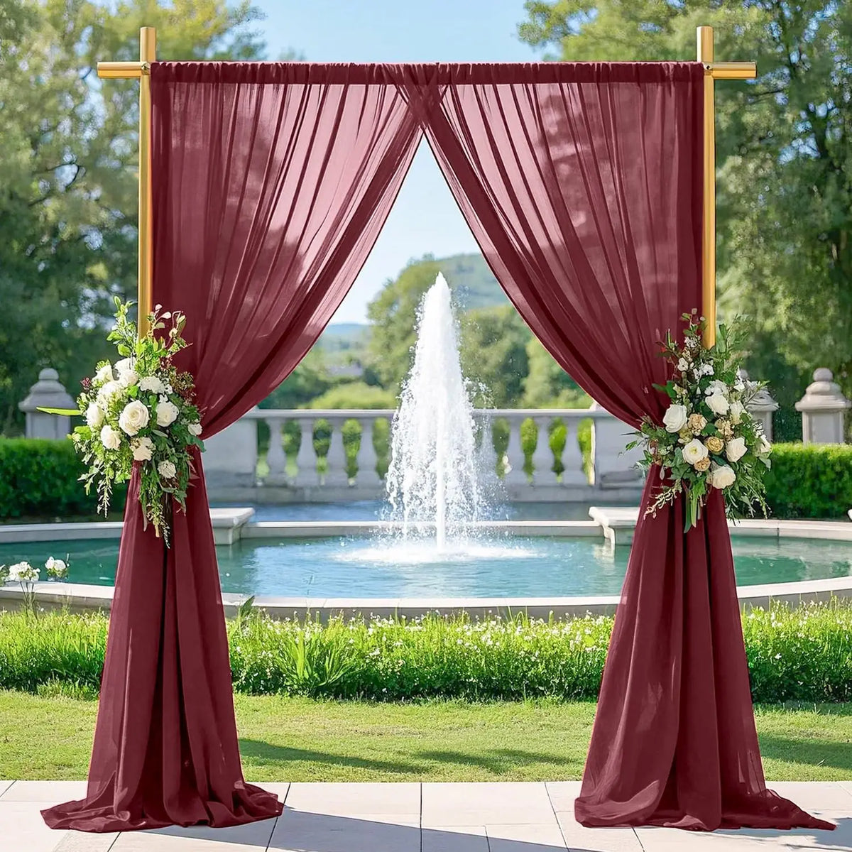 A maroon chiffon backdrop curtain draped in a wedding archway framing a fountain and green landscape.