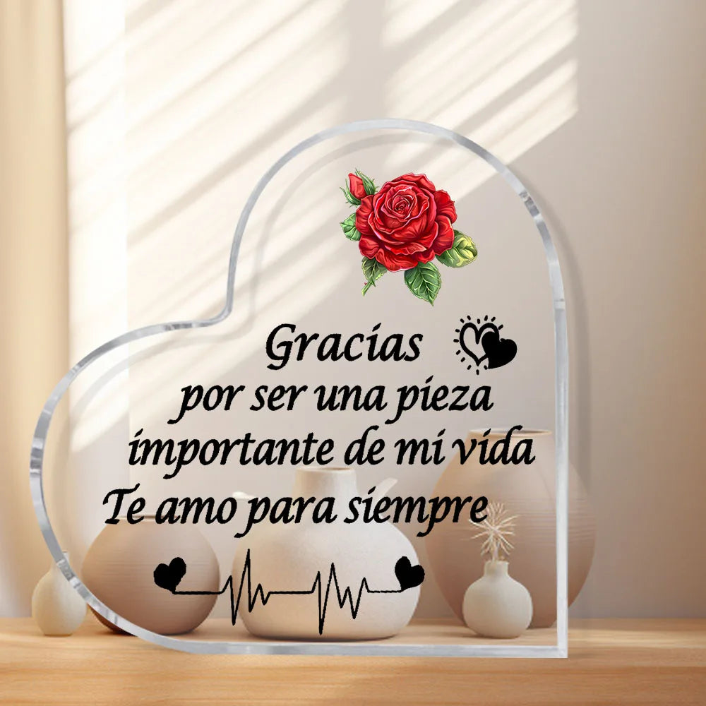A heart-shaped glass frame with a floral design and Spanish text inside, displayed on a wooden surface.