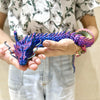A hand holds a purple and green articulated 3D printed dragon figurine.