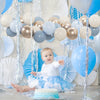 A baby sitting surrounded by blue and gold confetti balloons and white decor.