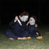Two individuals dressed as zombies with pale makeup and bloody knife props lying on grass at night