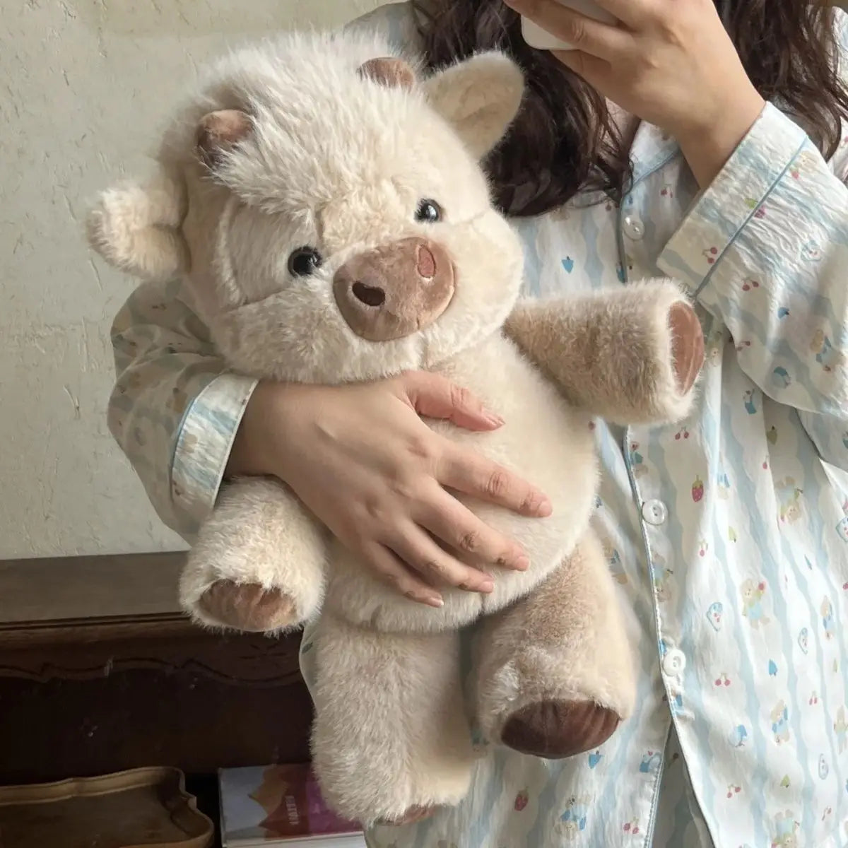 Une personne tient un jouet en peluche de veau écossais duveteux beige clair contre un t-shirt à motifs bleu clair.