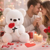 A white bear plush toy sits on a table surrounded by red roses, candles, and heart-shaped confetti.
