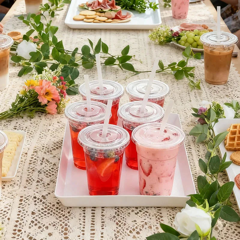 A pink and red fruit smoothie is served in a plastic cup on a white tray amidst greenery.