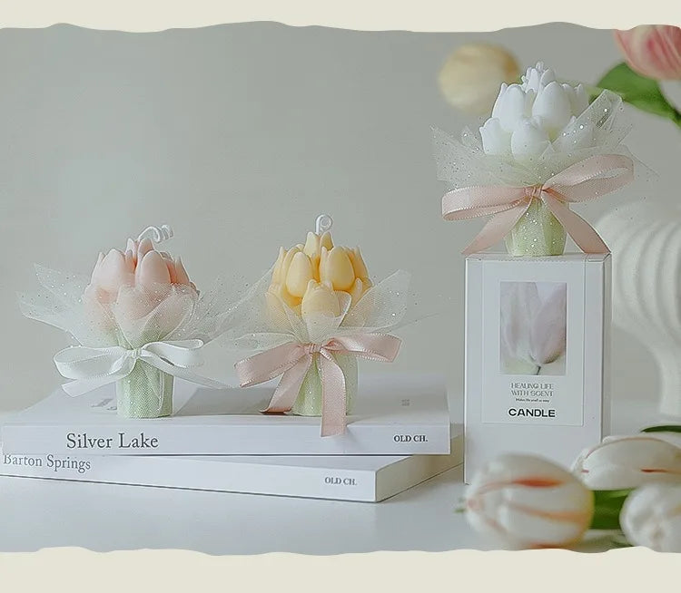 A white candle in a glass jar with a pink ribbon and a white tulip flower is displayed on a stack of books.