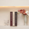 A set of three hardcover books with dark spines and white pages displayed on a light shelf next to a pink orchid arrangement.
