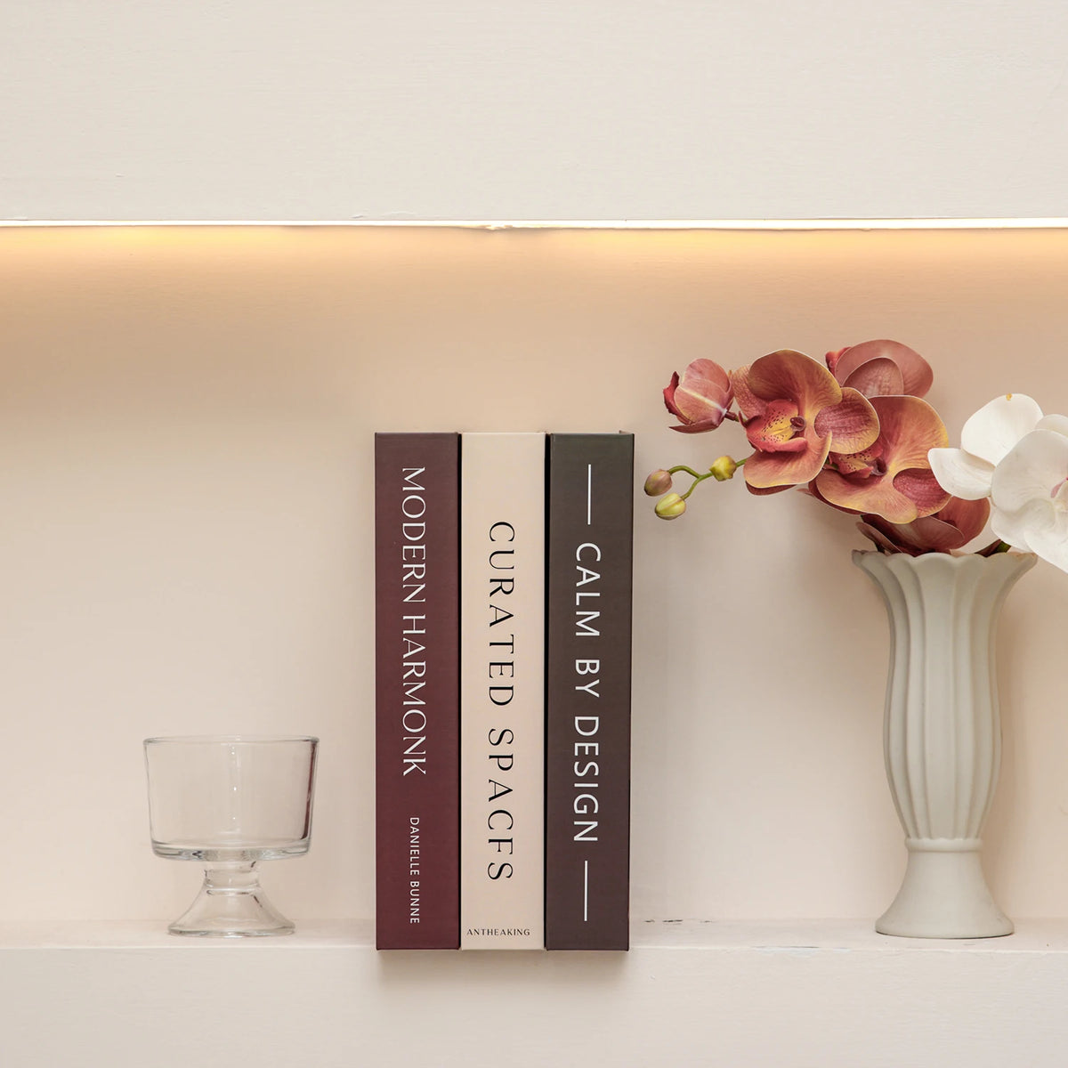 A set of three hardcover books with dark spines and white pages displayed on a light shelf next to a pink orchid arrangement.