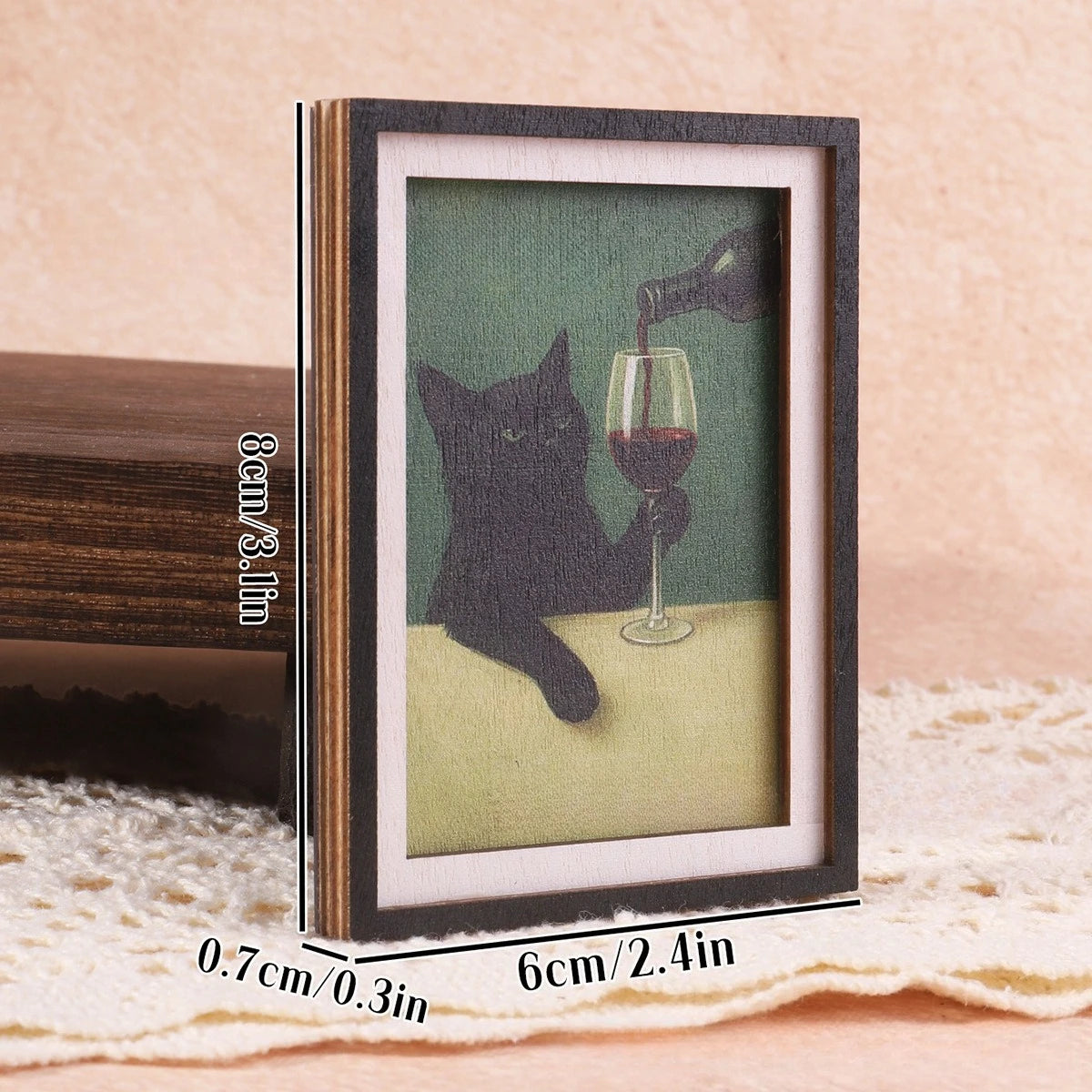 Gerahmtes Bild einer schwarzen Katze, die ein Glas Rotwein hält, auf einer hellen Oberfläche.