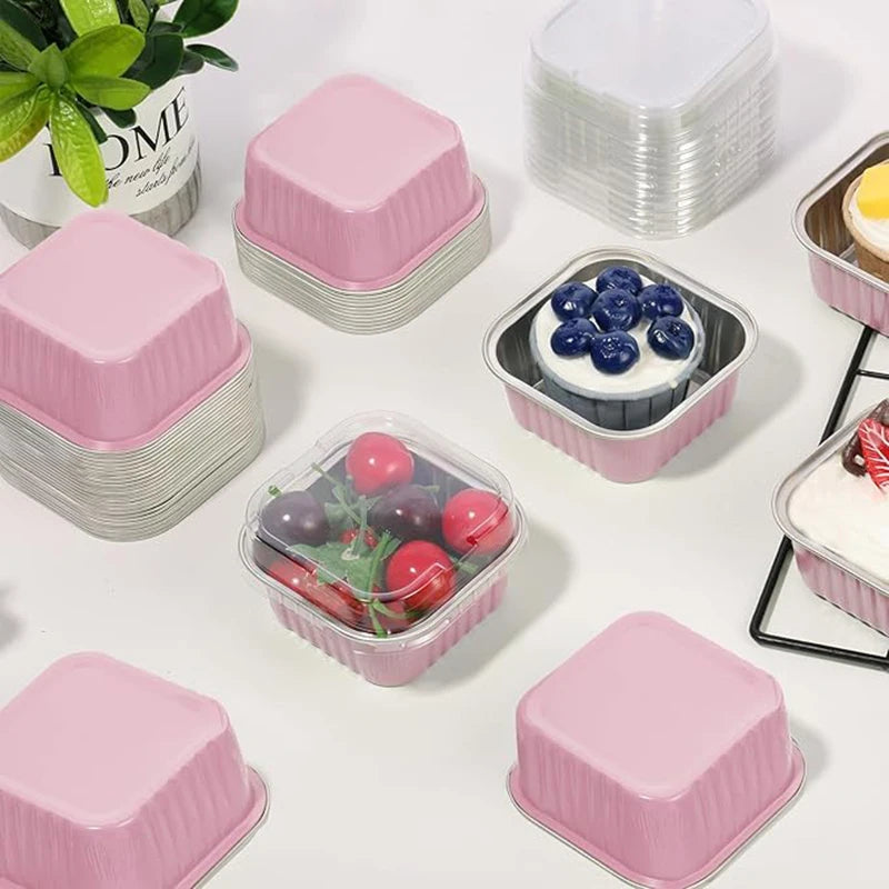 Pink reusable aluminum foil baking cups with lids and spoons arranged on a white surface.