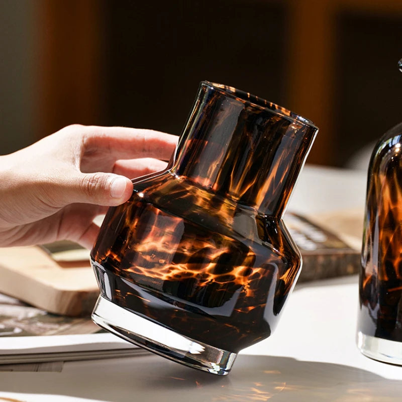 A hand holds a dark amber glass vase with an organic, mottled pattern against a light background.