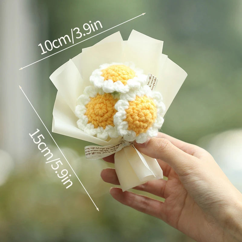 A hand holds a bouquet of handmade white and yellow crochet flowers wrapped in sheer paper.