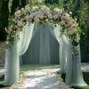 An elegant floral archway made of white roses and greenery draped with sheer light green tulle for an event backdrop.