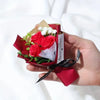 Une main tient une boîte cadeau rouge contenant des mini roses artificielles et une note manuscrite.