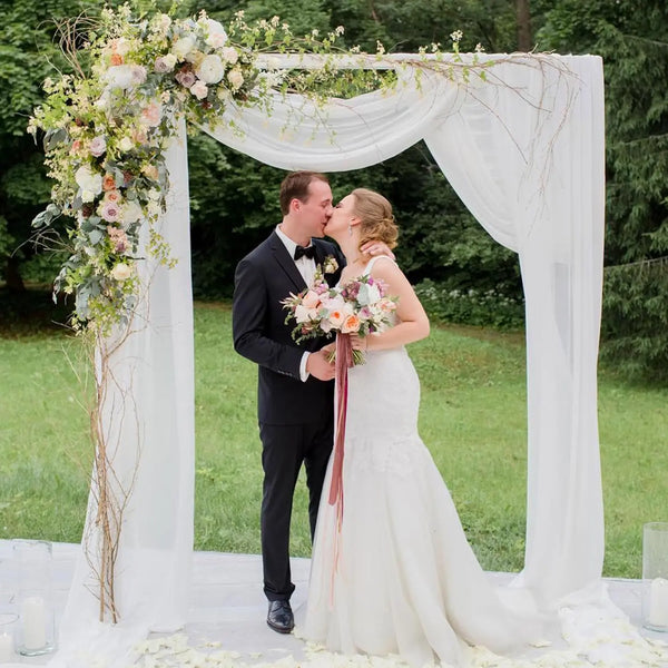 A bride and groom pose under a white sheer tulle draping fabric arch backdrop for a wedding photo.