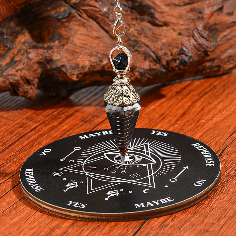 A silver pendant with a black crystal hangs from a gold-colored piece and rests on a wooden divination board.