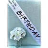 A white satin birthday sash with black text is displayed diagonally on a light blue fabric next to a white flower.