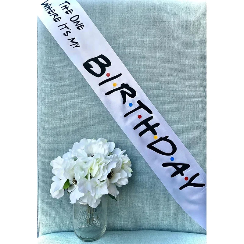 A white satin birthday sash with black text is displayed diagonally on a light blue fabric next to a white flower.