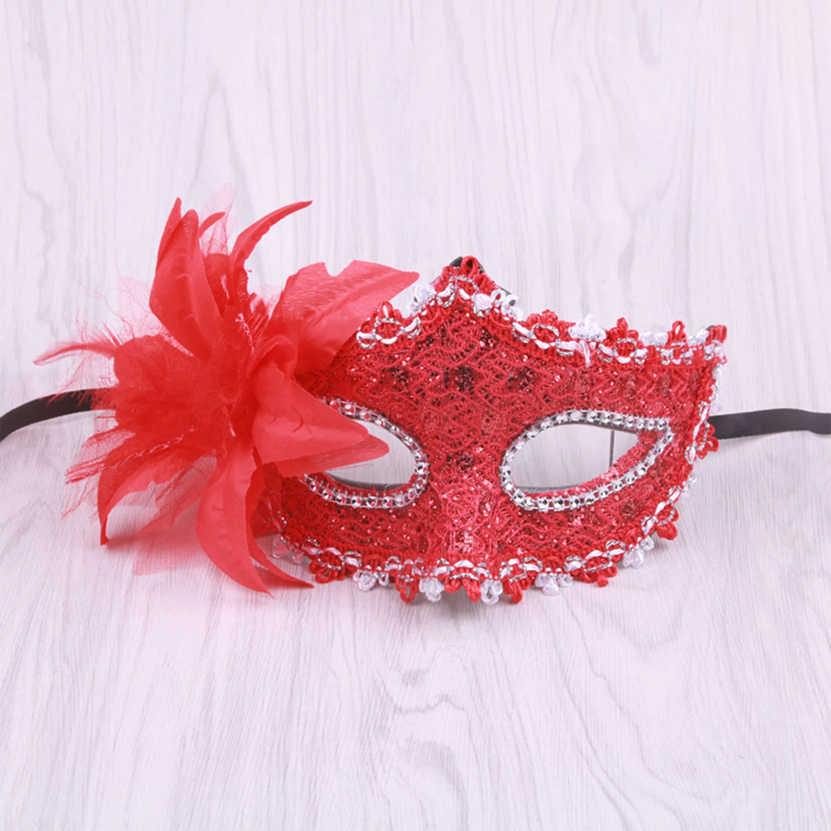 A red rhinestone masquerade mask with a feather accent displayed on a white wooden surface.