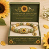 A gold and pearl stretch charm bracelet with a sunflower pendant displayed in a green box.