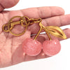 A gold-toned pendant keychain shaped like a pink glittery cherry leaf and fruit, held in a hand.