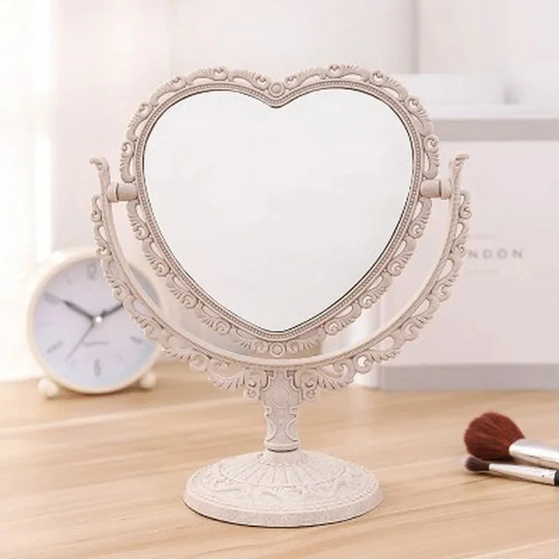 Espejo de maquillaje con forma de corazón y doble cara con marco blanco ornamentado sobre una superficie de madera.