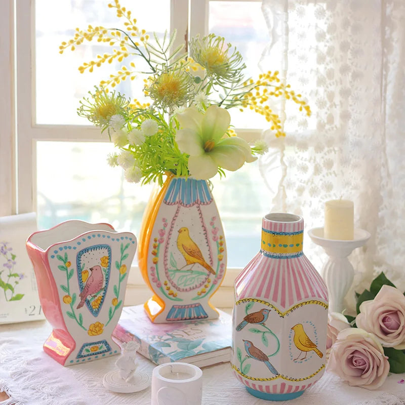 A ceramic bird floral vase with pink and yellow stripes sits on a white cloth near a window.