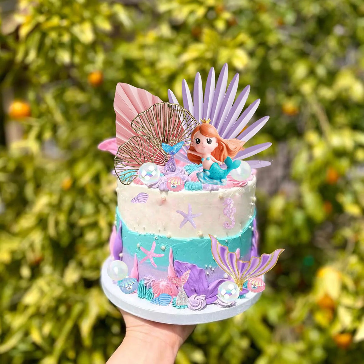 A tiered mermaid cake decorated with blue and pink frosting, featuring colorful sea elements like fins, shells, and seaweed.