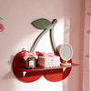 A red wooden shelf with a large green cherry-shaped cutout displaying a pink gift box and a silver mirror.