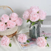 A bouquet of pink silk peony artificial flowers in a glass vase with a sheer ribbon and loose petals on a white surface.