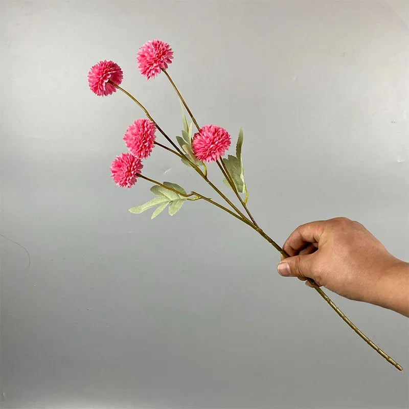 Una mano sostiene un tallo de flores artificiales de margarita de crisantemo rosa contra un fondo gris claro.