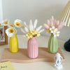 A pink ceramic bud vase holds white and yellow flowers and pampas grass on a wooden tray.