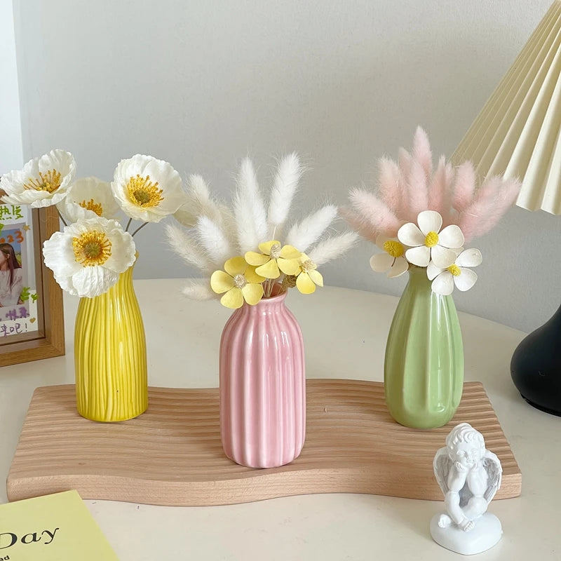 A pink ceramic bud vase holds white and yellow flowers and pampas grass on a wooden tray.