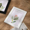 A white picture frame displaying a green plant illustration on a wooden surface.