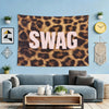 A large brown leopard print tapestry wall hanging featuring the word SWAG in light pink text, displayed above a blue wall.