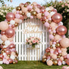 A large balloon garland archway featuring pink, gold, and white balloons set against a white wooden fence.
