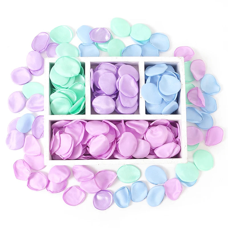 Una colección de pétalos de flores de seda artificial en varios tonos de rosa y azul dispuestos alrededor de una caja blanca.