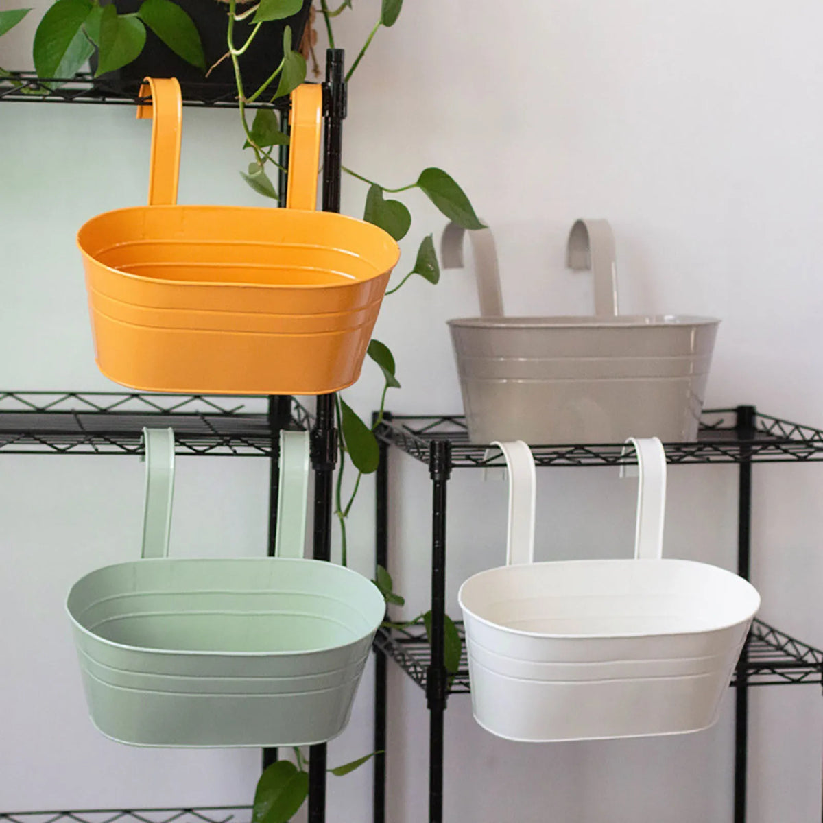 Three modern metal hanging planters in orange, gray, and white are displayed on a black wire rack.