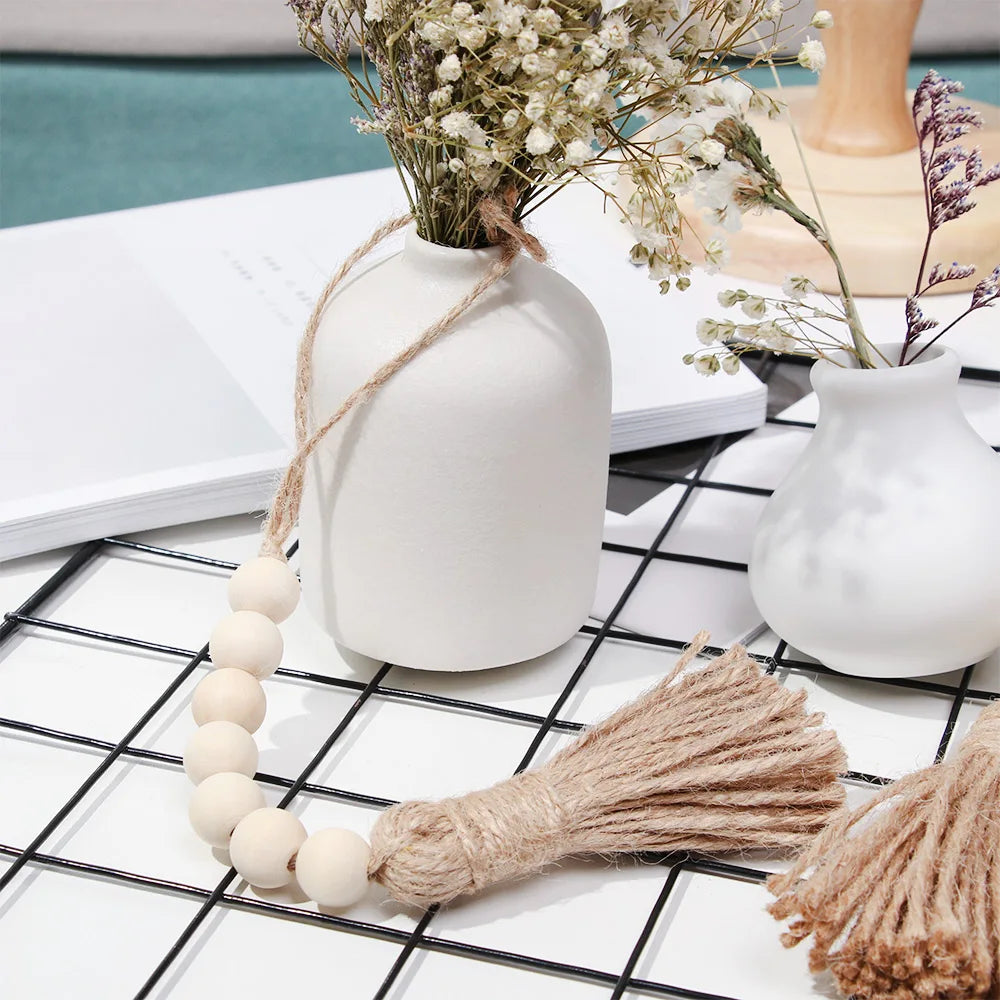 A handmade wood and jute bead garland displayed next to a white ceramic vase with dried flowers on a black grid surface.