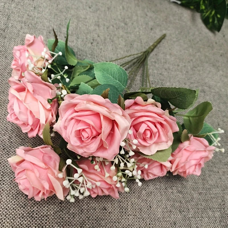 A bouquet of pink roses and green foliage with small white filler flowers on a gray fabric background.